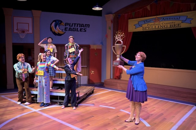 Cast members perform a scene from The 25th Annual Putnam County Spelling Bee on a colorful stage set as a school gym, while a woman holds up a large trophy and the students celebrate behind her.