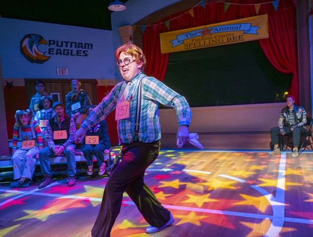 A spelling bee contestant wearing number 157 dances energetically across a brightly lit gymnasium stage while other contestants watch from the bleachers during The 25th Annual Putnam County Spelling Bee.